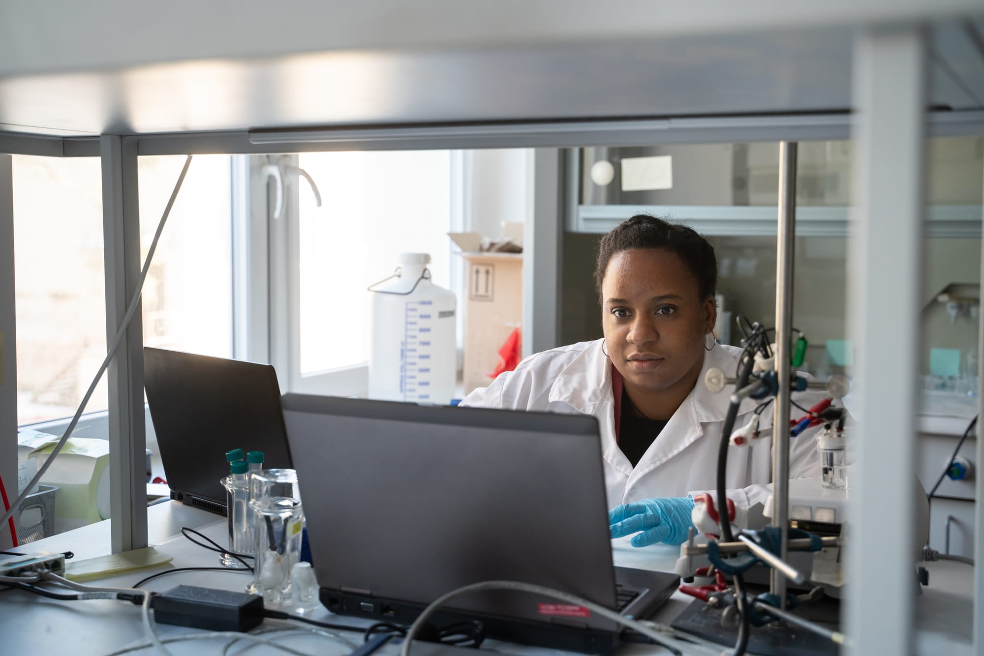 Pesquisadora concentrada analisando dados em um laptop ao lado de equipamentos de laboratório, sob luz natural.